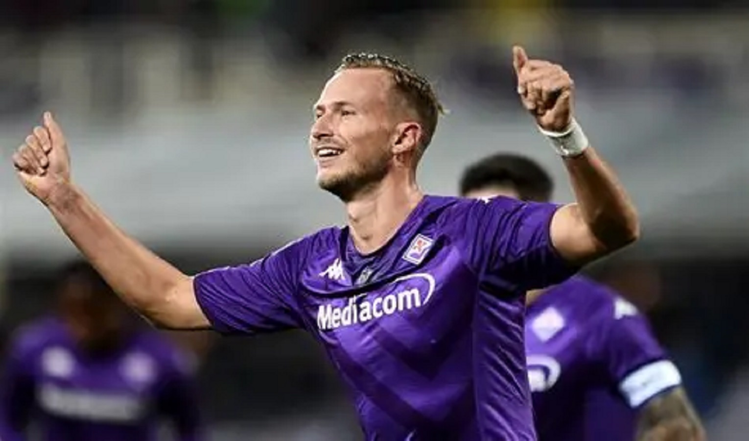 Un calciatore in maglia viola sorride e allarga le braccia per festeggiare