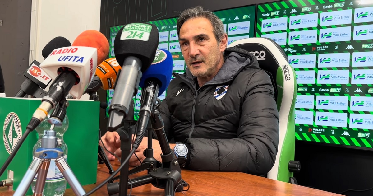 Un uomo seduto che parla a una conferenza coi microfoni davanti