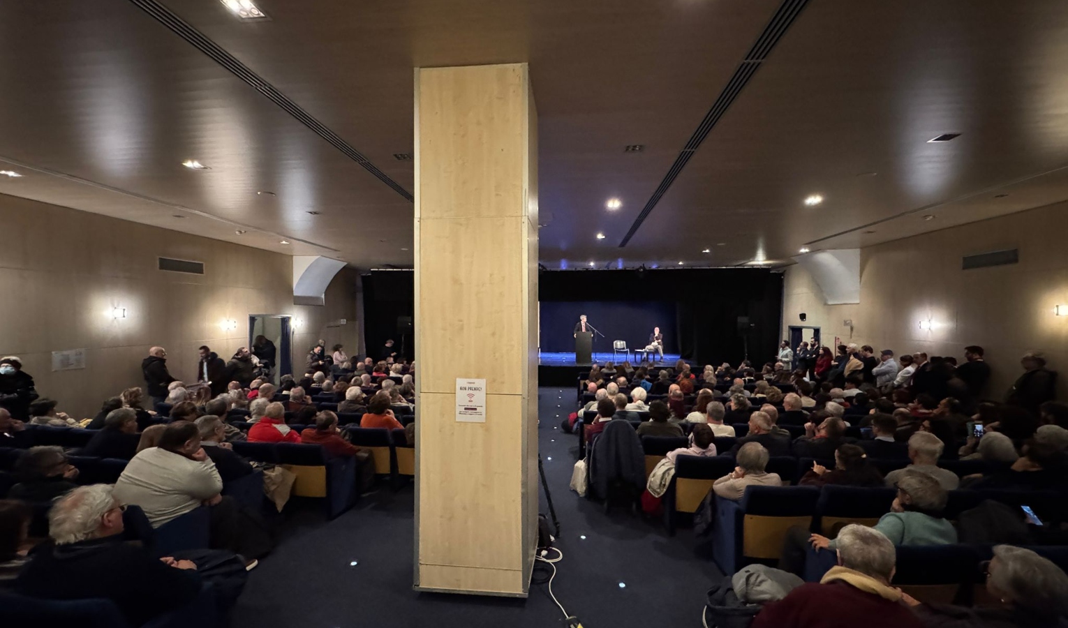 Gente seduta in sala e persona in piedi che parla dal palco