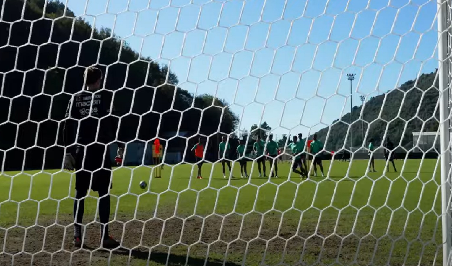 Un portiere di calcio davanti alla porta, di schiena. In corso un allenamento su un campo di calcio con altri calciatori